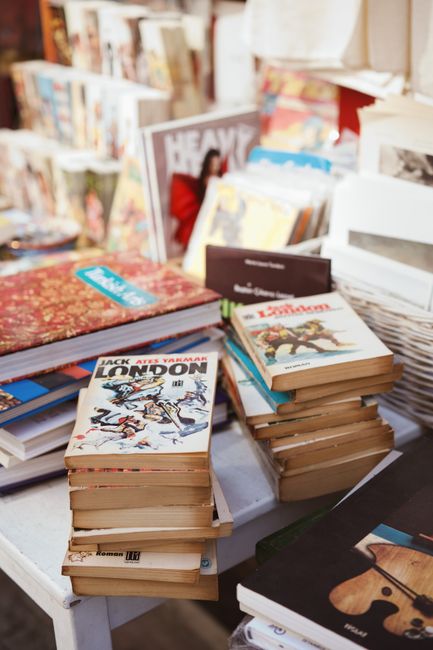 A detailed close-up view of several stacks of paperback books placed on a white surface, with some overlapping others. The books feature various cover designs, with one prominently displaying an illus