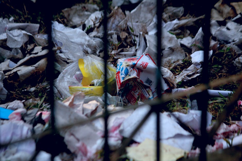 A large red metal skip with visible rust and chipped paint sits against a dark green wall on a paved outdoor area, with a light gray concrete wall to its right. Several white plastic bags filled with