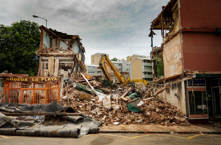 A pile of household waste and debris on the pavement beside a building exterior, including rolled-up rugs with intricate multicoloured patterns, wooden slats and broken furniture parts, and several la