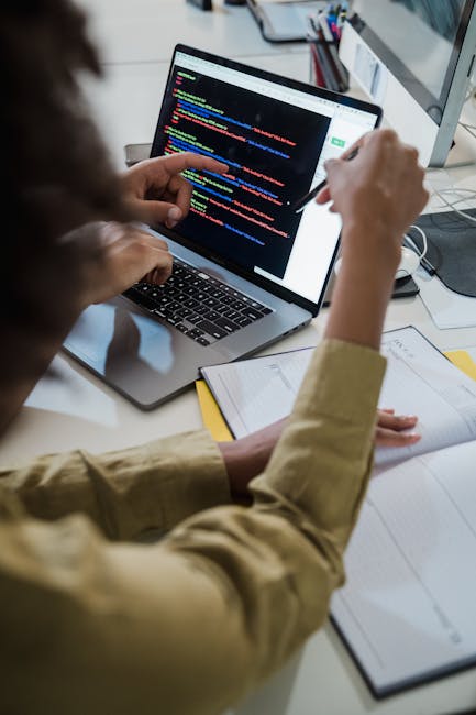 A person working at a desk, viewed from behind, interacting with a laptop displaying a code editor with multiple lines of colored programming code on the screen. The individual is wearing a beige long