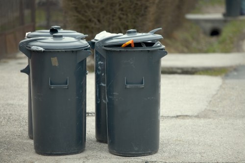 Two dark grey plastic wheelie bins with hinged lids are positioned on a paved driveway or street surface, with one lid slightly open revealing an orange-handled tool or object inside. The bins are situated in a residential outdoor setting, with blurred greenery and parts of a house visible in the background. The surface appears to be concrete or tarmac, and the bins are standard size for rubbish collection. The scene suggests a domestic environment where rubbish or waste collection is taking place, reflecting typical disposal practices. Property Clearance London provides rubbish removal services that could include the collection and disposal of such waste containers and their contents, especially in the context of property or garden clearance activities in London or surrounding areas. The lighting indicates daytime, with natural light illuminating the scene evenly, revealing the textures and colors of the bins and their environment clearly.