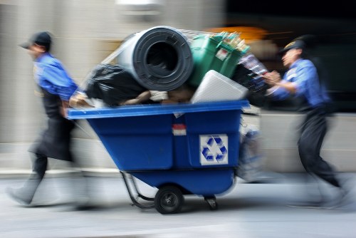 A blue wheeled rubbish bin is shown overflowing with various waste items, including a large black plastic bag, a green recycling bin, and a white container. Two individuals in blue uniforms are pushing the bin along a pavement or street; their motion is blurred, indicating movement. The background features a neutral-colored wall or building typical of urban environments, with the scene suggesting a waste collection or rubbish removal activity consistent with professional services in London or surrounding areas. The image emphasizes the handling of mixed waste materials in a professional removal setting, aligned with property clearance and rubbish disposal services provided by Property Clearance London.