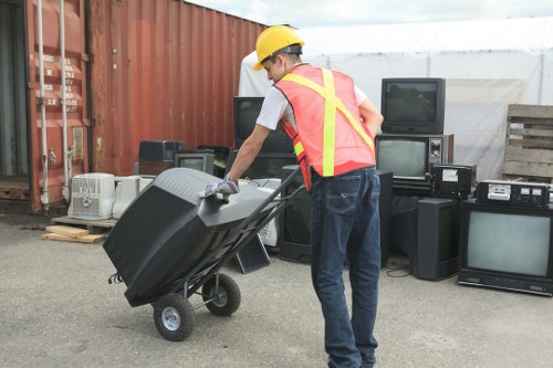 A waste collection worker wearing a yellow safety helmet and a high-visibility vest is pushing a large black plastic bin on wheels, likely filled with rubbish or discarded items, across an outdoor area adjacent to a row of old, used electronic appliances including televisions and stereo systems. The background includes a red corrugated metal fence, a pallet with additional items, and a partly covered structure with a white tarp or canopy. The ground appears to be paved, and natural daylight illuminates the scene, suggesting a typical rubbish removal operation possibly in or near London. Property Clearance London’s service appears relevant to this context, focusing on the collection and disposal of various waste materials, including electronic waste, in the local area. The scene captures the practical aspect of rubbish removal, emphasizing the handling of bulky refuse in an outdoor industrial or commercial setting, with attention to safety gear and relevant environmental management.